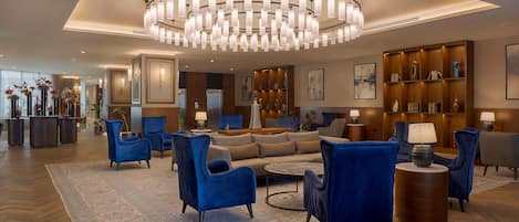A spacious lobby with a large chandelier, blue armchairs, a sofa, and a wooden bookshelf.