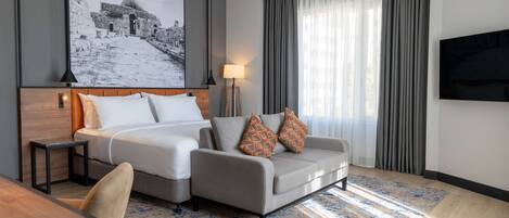 A modern hotel room with a bed, a sofa, a desk, a lamp, a TV, and a large framed picture on the wall.