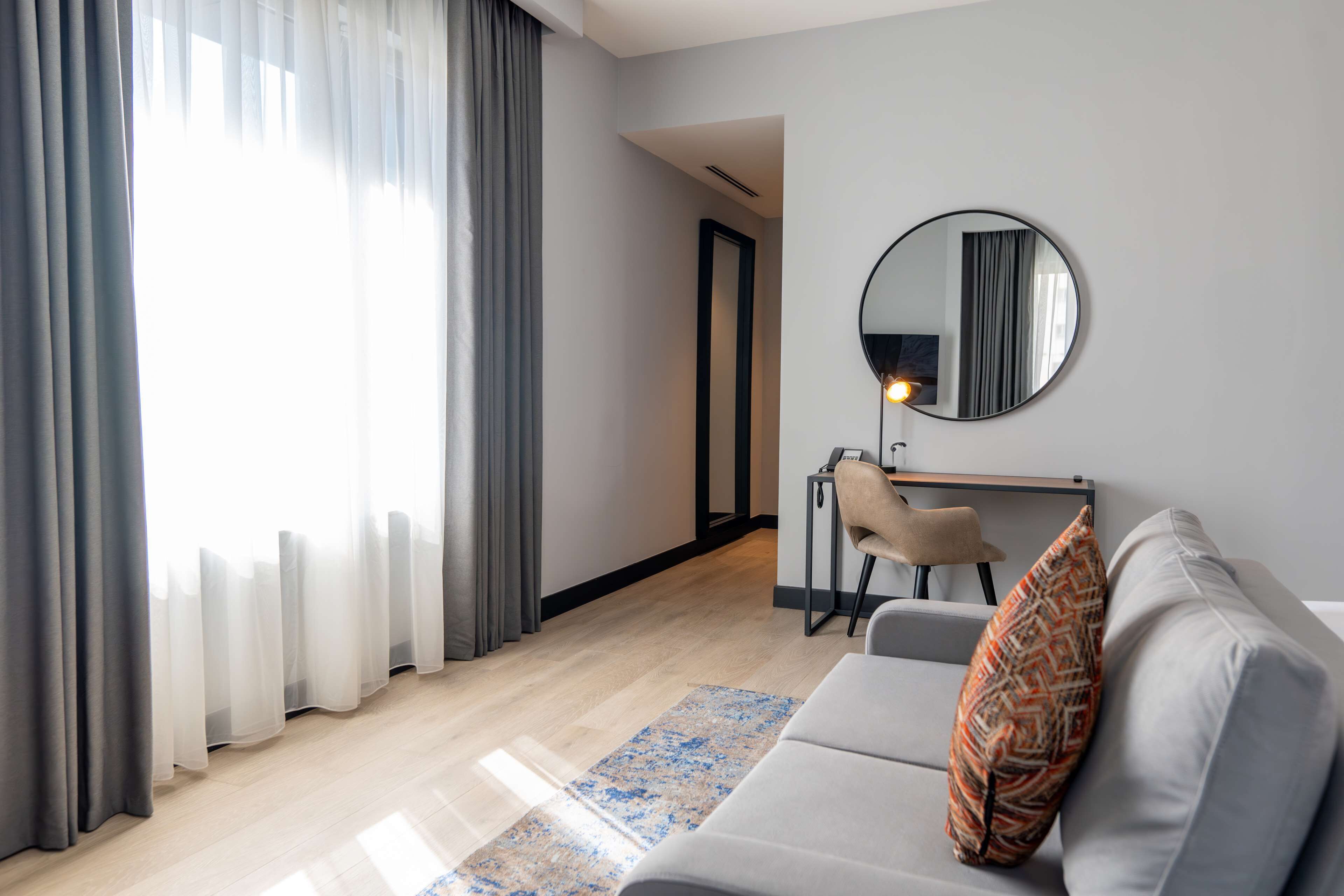 A modern living room with a grey sofa, a patterned throw pillow, a round mirror on the wall, and a view through sheer curtains to a bright window.