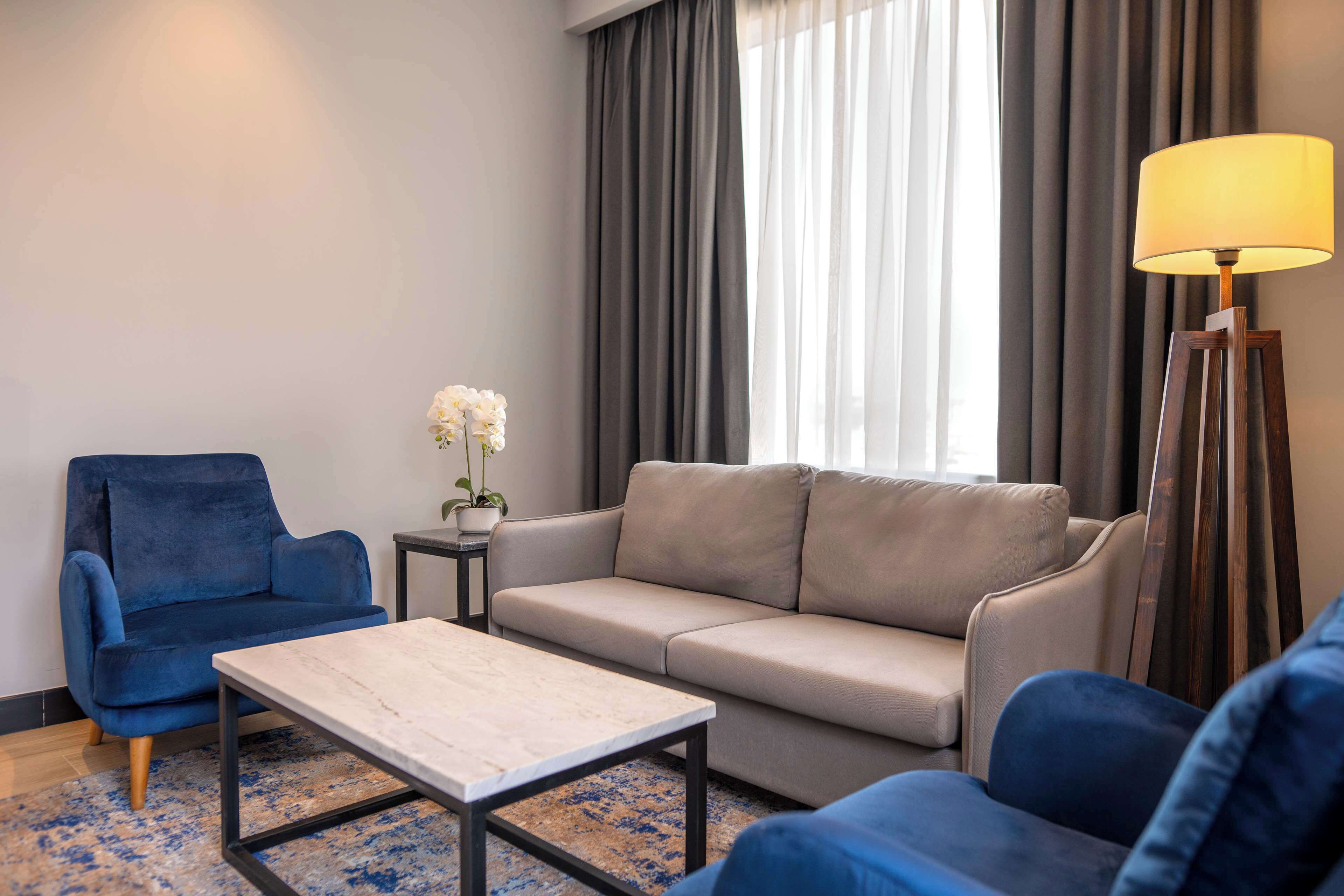 A modern living room with a blue armchair, a beige sofa, and a wooden coffee table. A floor lamp and a potted plant are also visible.