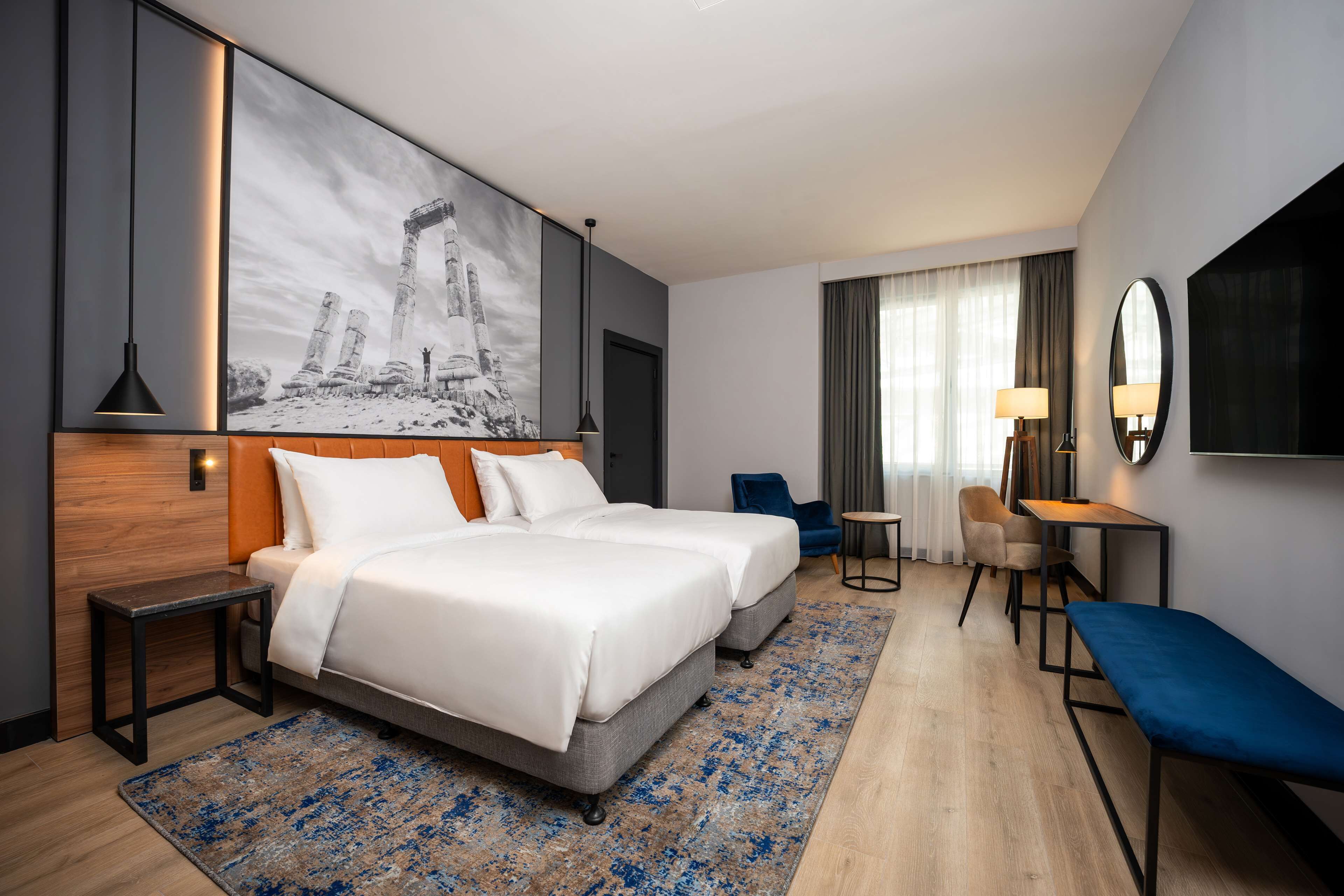 A hotel room with two beds, a desk, a chair, a TV, and a large painting on the wall.