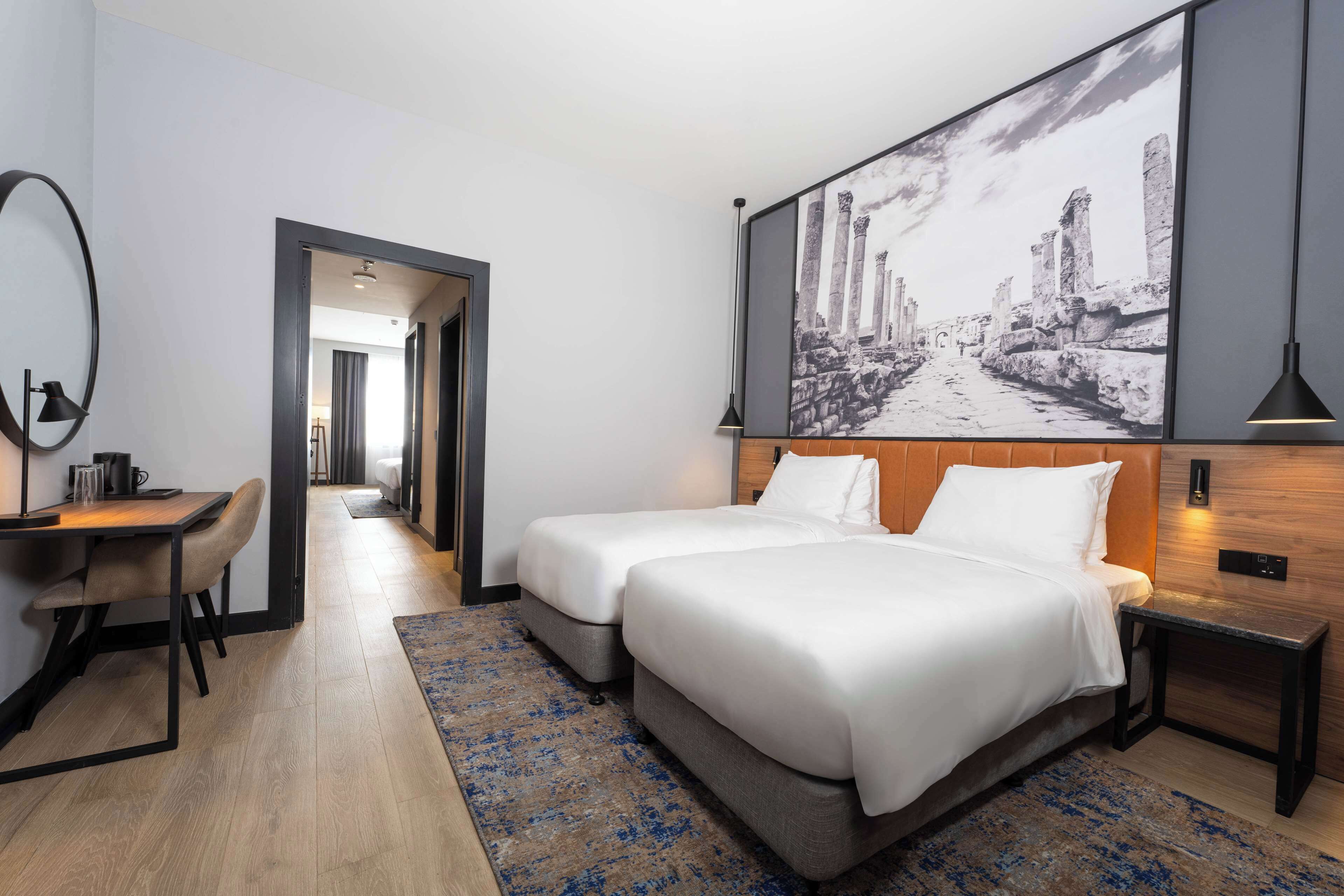 Quarto de hotel com duas camas, uma escrivaninha e um quadro grande na parede.