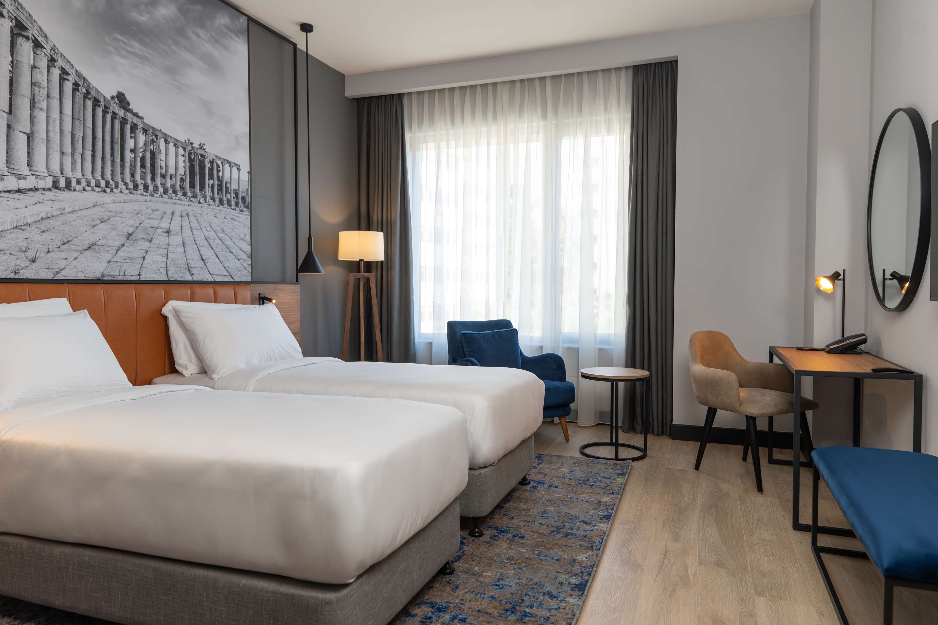 Hotellihuone, jossa on kaksi sänkyä, työpöytä, tuoli, pieni pöytä, lamppu ja suuri ikkuna, jossa on verhot.