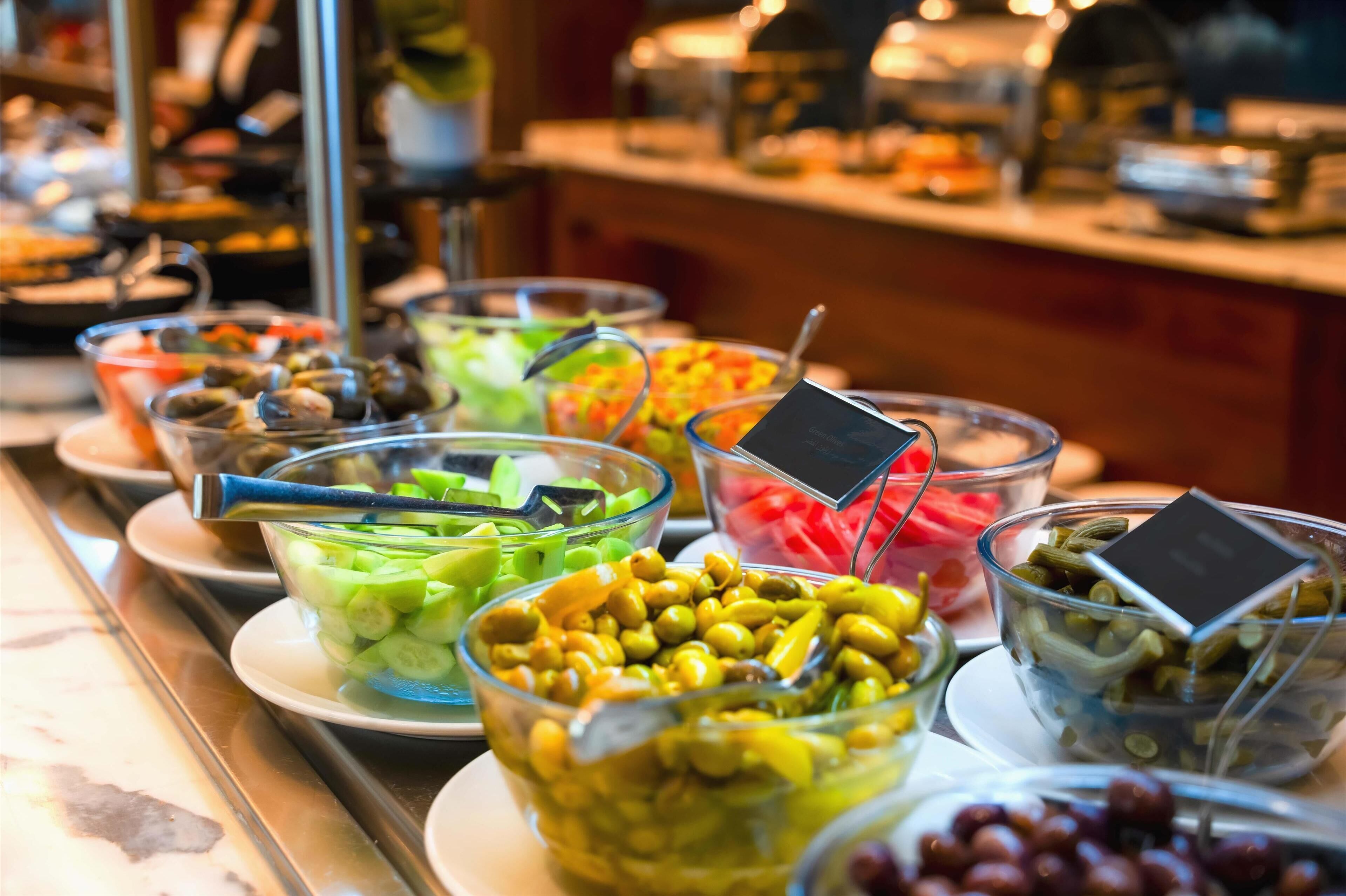A buffet with various food options including olives, vegetables, and fruits.