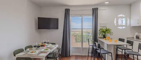 Una zona de menjador amb una taula preparada per a quatre, un televisor fixat a la paret i vista a l'exterior a través d'una porta corredissa.