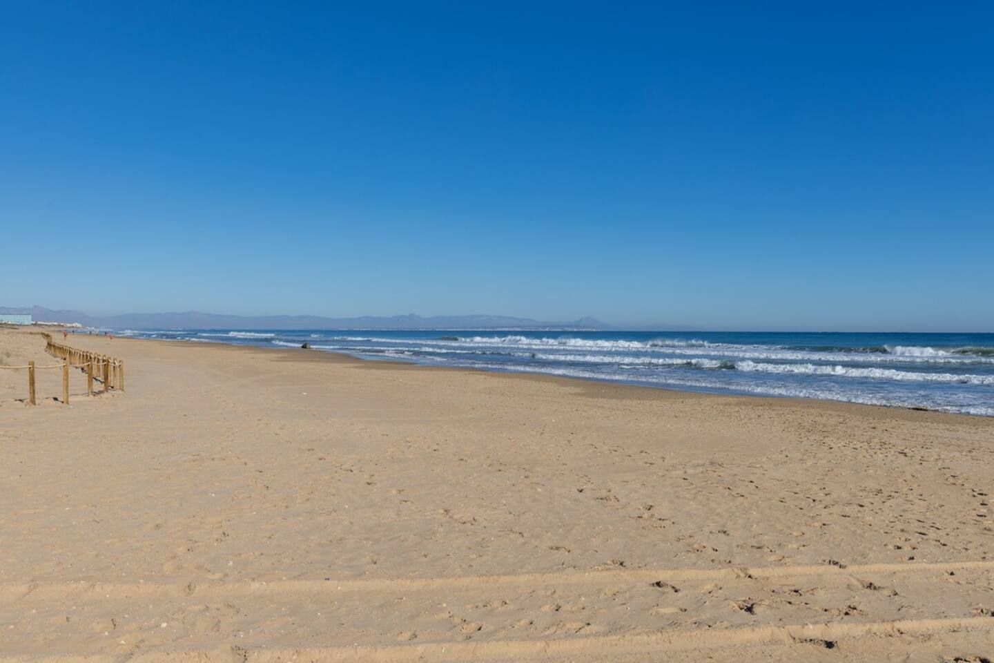 Une plage de sable avec une promenade en bois menant à l’eau, un ciel dégagé et des montagnes à l’horizon.