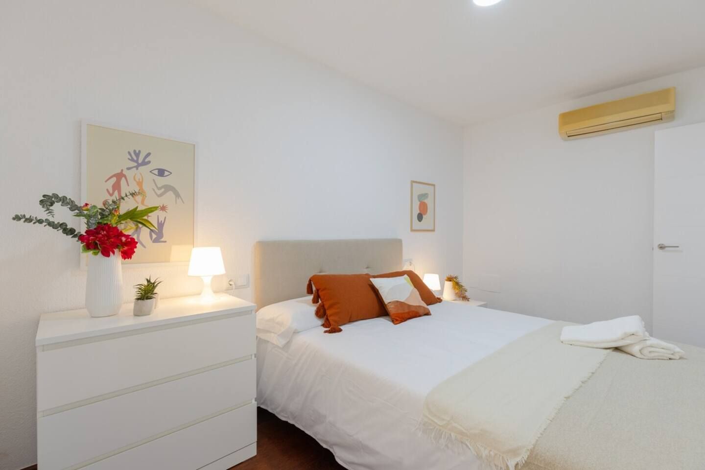 A hotel room with a bed, bedside tables, a nightstand, a wall-mounted air conditioner, and artwork on the walls.