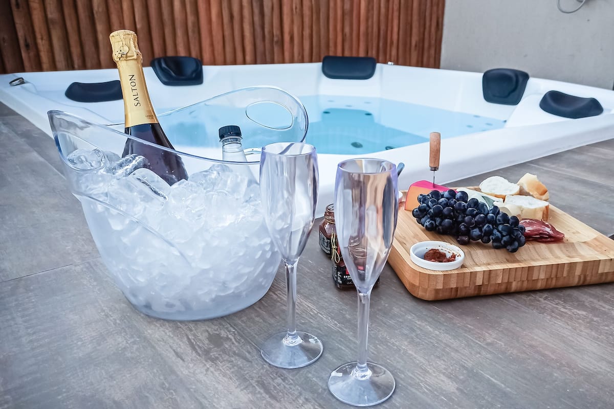 Ein Whirlpool mit Holzterrasse, eine Champagnerflasche, mit Eis gefüllte Gläser und ein Holzbrett mit Trauben und Käse.