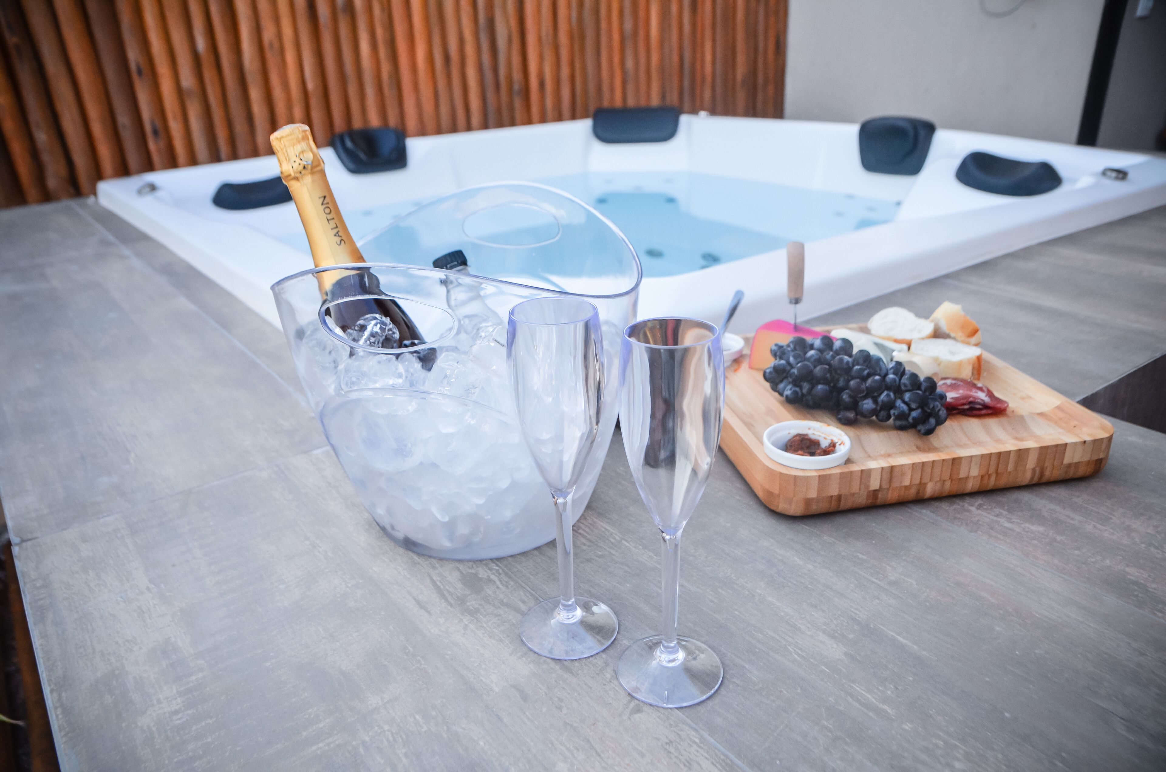 Un jacuzzi con champán y copas sobre una mesa, y una tabla de madera con uvas y pan.