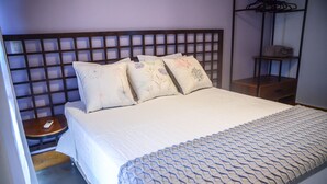 Una cama bien hecha con un edredón estampado y almohadones decorativos, una mesita de noche con un control remoto y un buró de madera con un estante.