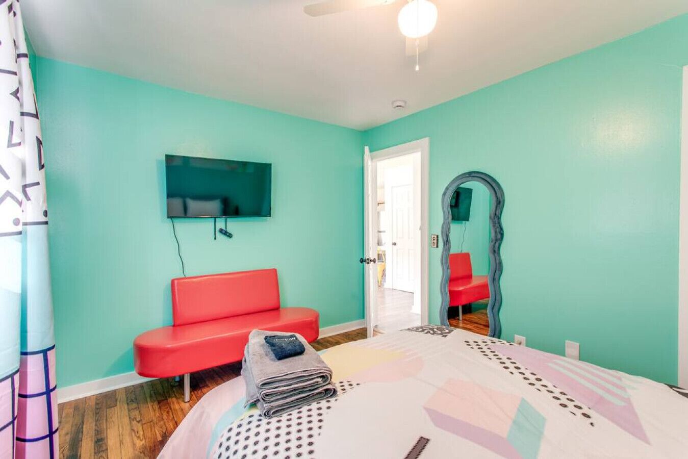Una habitación con paredes color turquesa, un sofá rojo, una cama con edredón estampado, un ventilador de techo y un televisor montado en la pared.