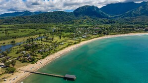 Vista aérea de um resort costeiro com uma praia, um píer e montanhas verdejantes ao fundo.