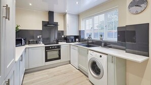 A modern kitchen with white cabinetry, a built-in oven, a washing machine, a microwave, and a large window with blinds.