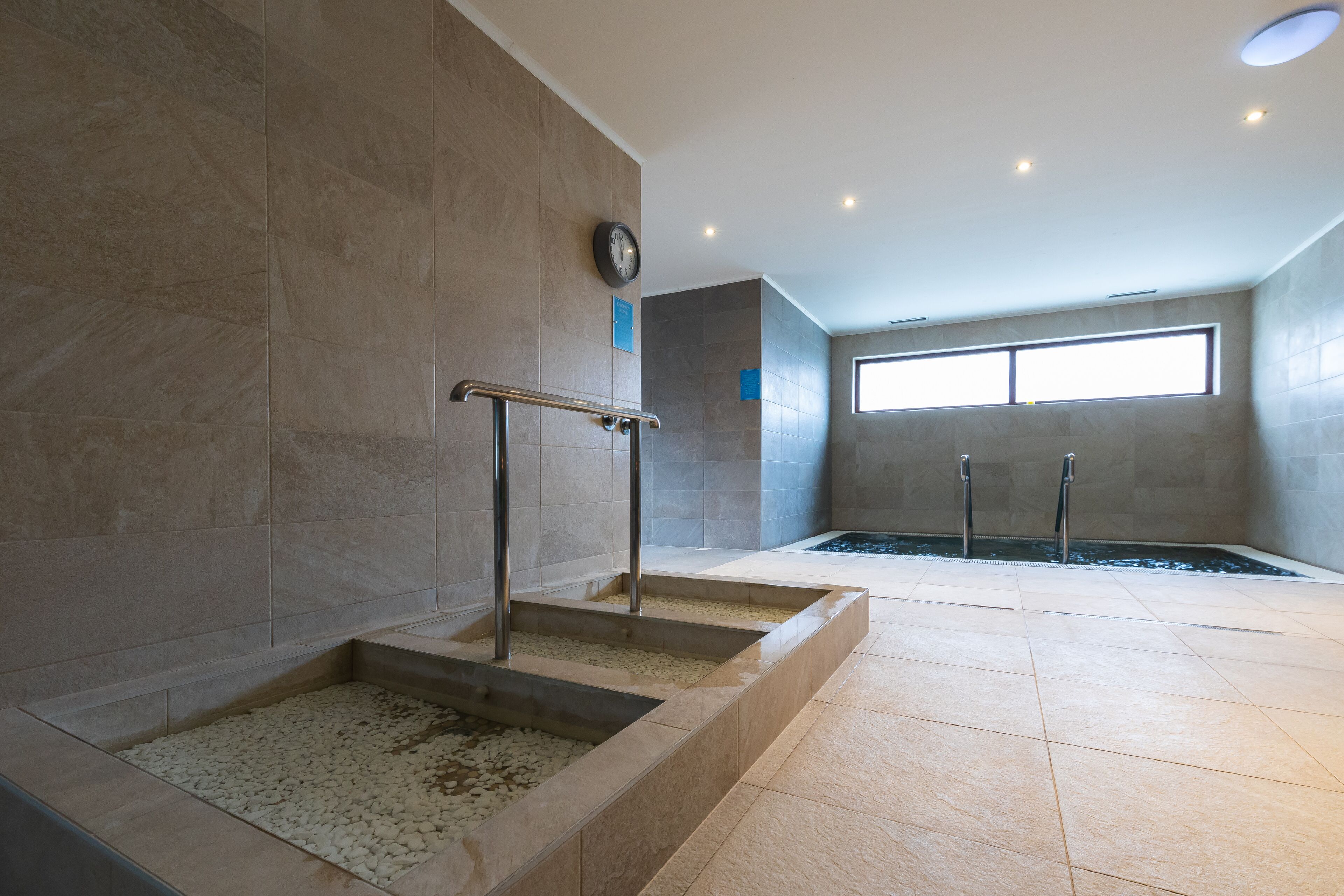 Un área de piscina interior moderna con paredes de azulejos, un reloj y un ventanal que proporciona luz natural.