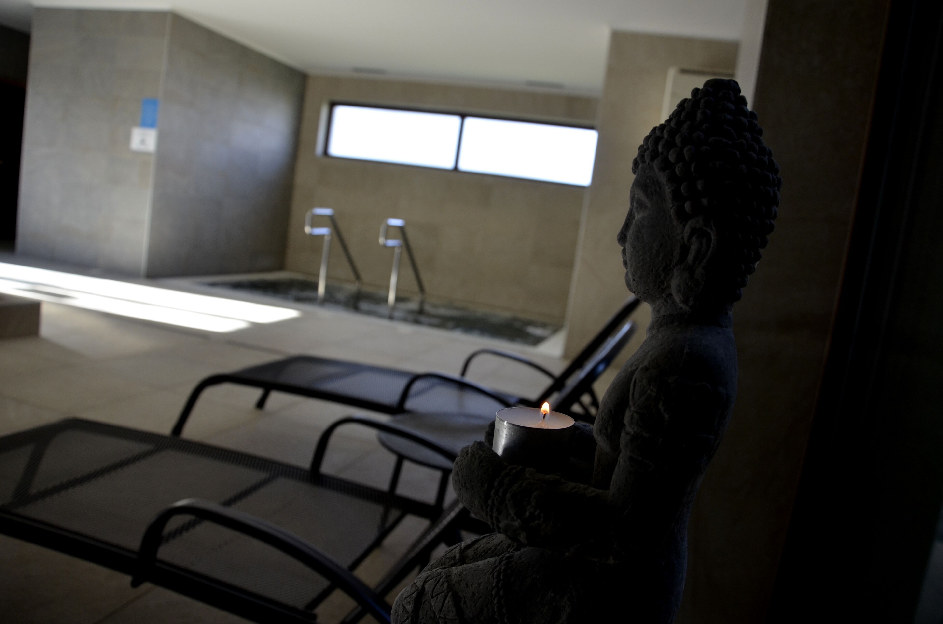 Eine Buddha-Statue hält eine Kerze in einem schummrigen Raum mit Fenster und Pool im Hintergrund.