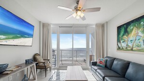A living room with a large TV, a ceiling fan, a sofa, a coffee table, and a view of the beach through the glass doors. - Lighthouse 711 by Liquid Life (Gulf Shores)