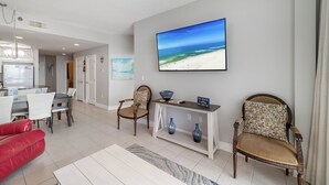 A spacious living room with a large flat-screen TV, comfortable seating, a dining area, and a kitchen in the background. - Lighthouse 711 by Liquid Life (Gulf Shores)