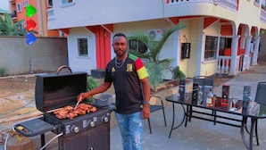 Un hombre asando al aire libre con una barbacoa, una mesa con bebidas y un edificio al fondo.