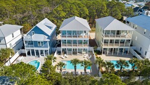 A coastal property with multiple buildings, a large swimming pool, and a forested backdrop. - Seagrove - 1 Block from Beach, Golf Cart, Private HEATED Pool, Game Room, Bikes & Tesla Charger! (Santa Rosa Beach)
