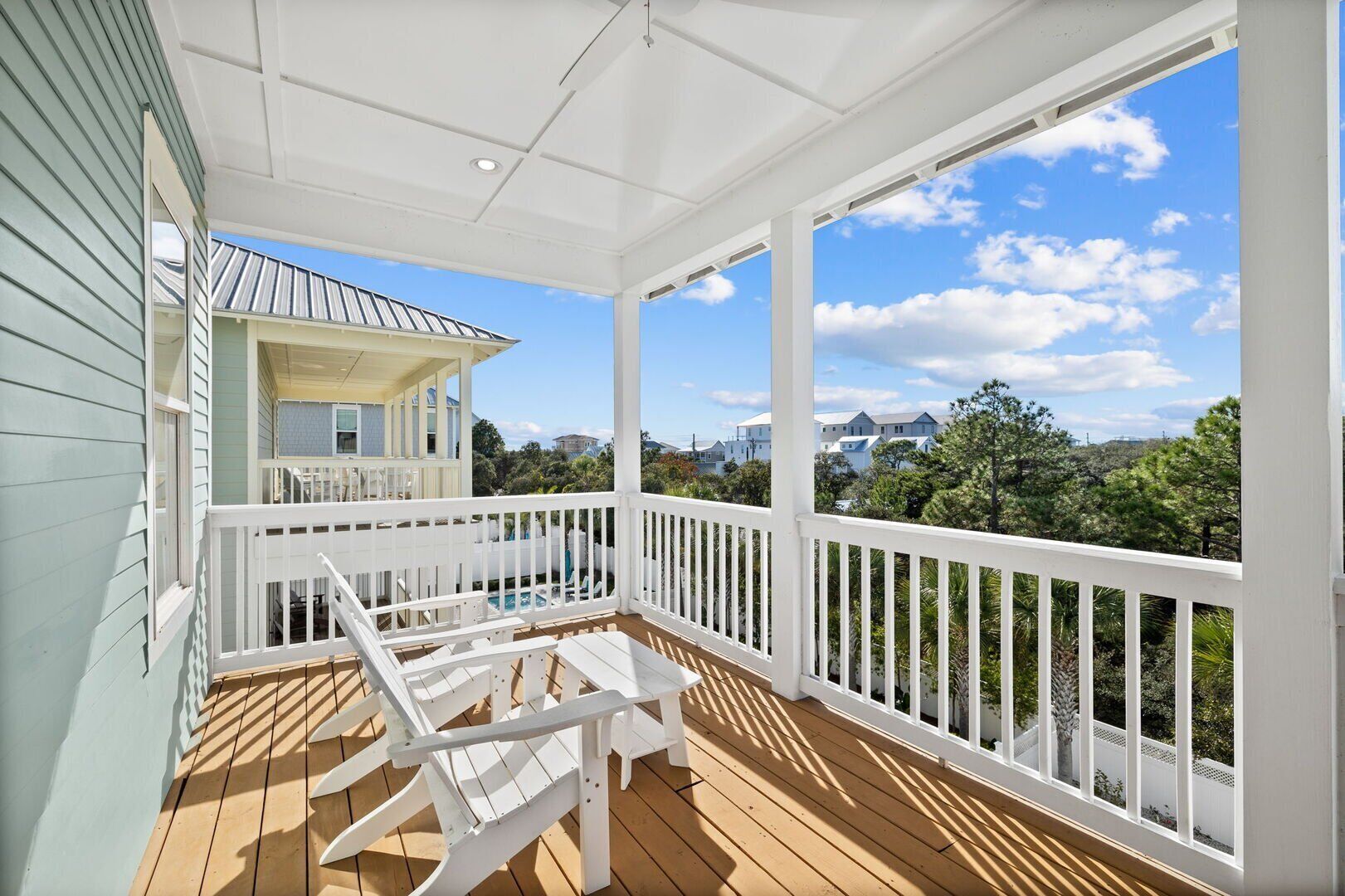 A covered outdoor deck with white railings and wooden flooring, overlooking a residential area with houses and trees.