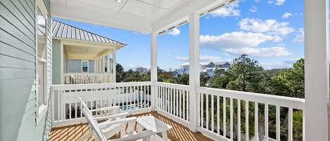 A covered outdoor deck with white railings and wooden flooring, overlooking a residential area with houses and trees.