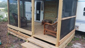 Een houten terras met een overdekte veranda, rieten meubilair en een schuifdeur die toegang geeft tot een voertuig.