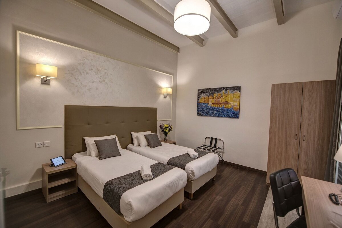 A hotel room with two beds, a desk, a chair, a painting, and a wardrobe.