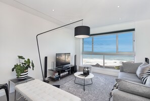 A modern living room with a large window overlooking the ocean, a flat-screen TV, a coffee table, and a sofa. - Blouberg Heights 406 (Cape Town)