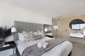 A modern hotel room with a large bed, a bedside table with a lamp, a round mirror, and a built-in vanity with a sink. - Blouberg Heights 406 (Cape Town)