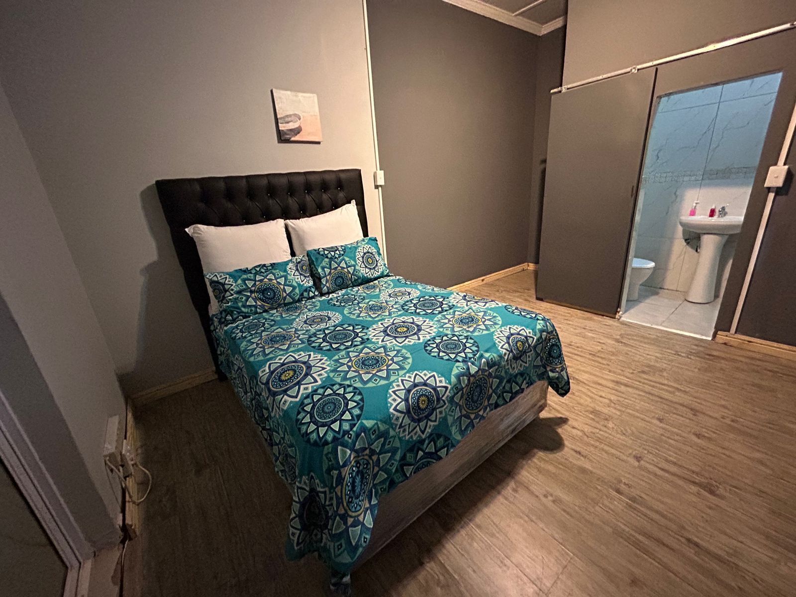 Une chambre à coucher avec un lit, une tête de lit, un tableau accroché au mur et une salle de bain visible à travers une porte en verre.