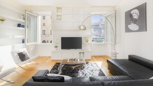 A modern living room with a large flat-screen TV, a black leather sofa, a glass coffee table, and a herringbone-patterned rug.