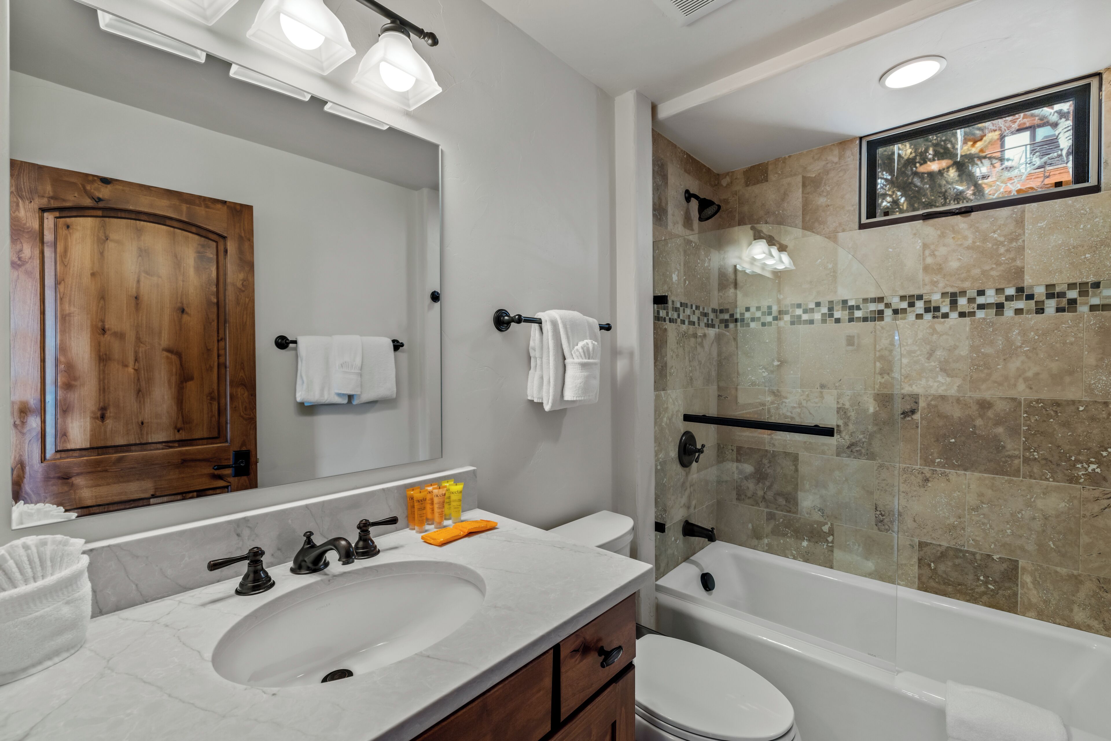 A modern bathroom with a double sink, a large mirror, a wooden door, a shower with a glass enclosure, and a view of the outside through a window.