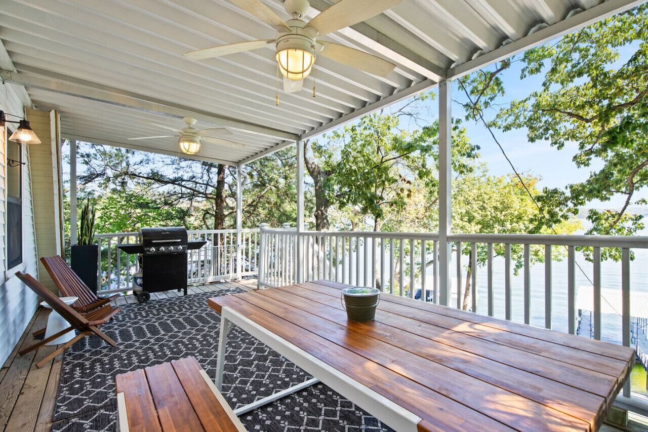 A covered outdoor area with a wooden table, chairs, a grill, and a view of the water.