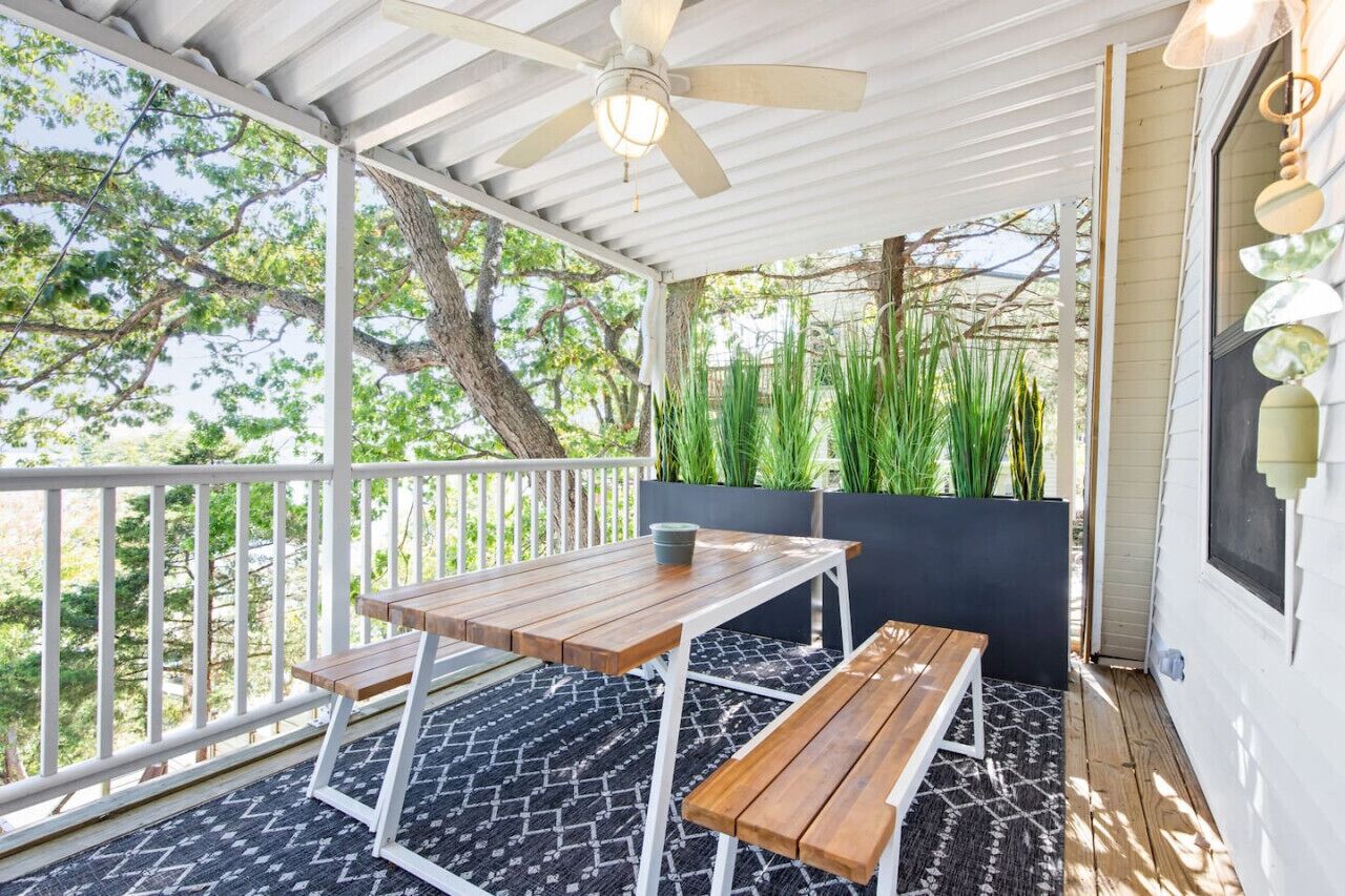 A covered outdoor patio with a table and benches, surrounded by greenery and trees.