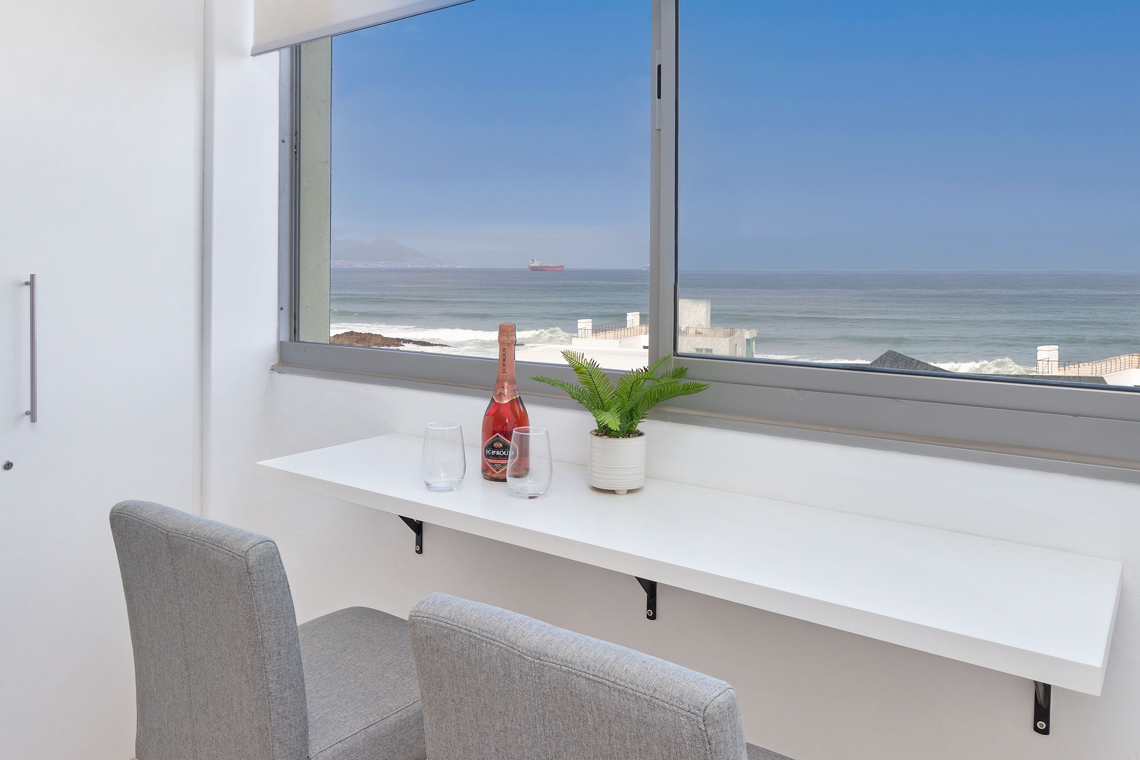 A modern room with a large window overlooking the ocean, a white bench, two grey chairs, a small potted plant, and a bottle of wine.
