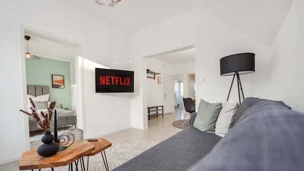 Un salon moderne avec un canapé gris, une table basse en bois et un téléviseur fixé au mur qui affiche le logo « NETFLIX ».