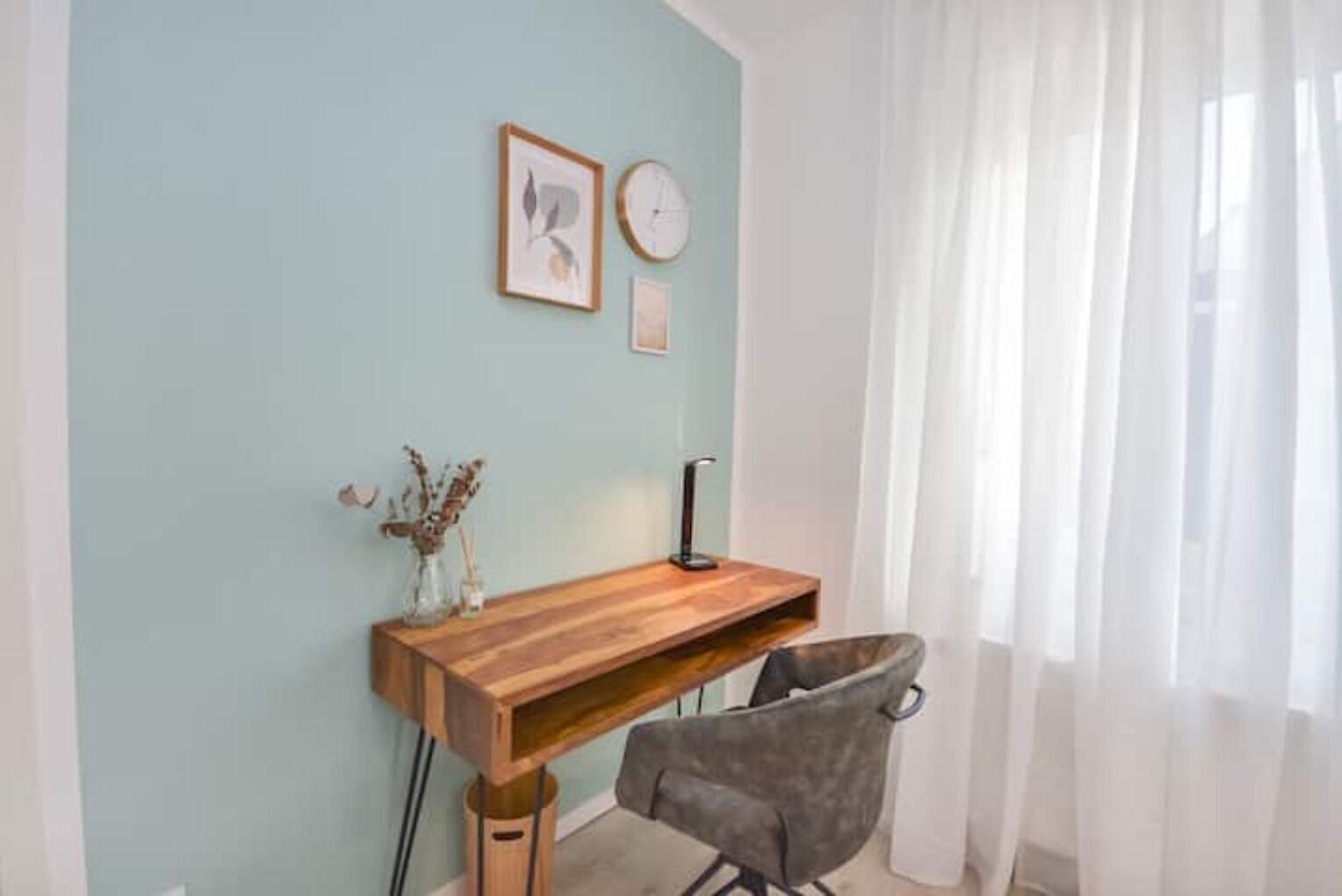 Una habitación con un escritorio de madera, una silla gris, un jarrón con flores secas y un reloj en la pared.