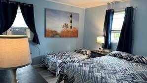 Une chambre avec deux lits, un tableau représentant un phare, une lampe et des rideaux.