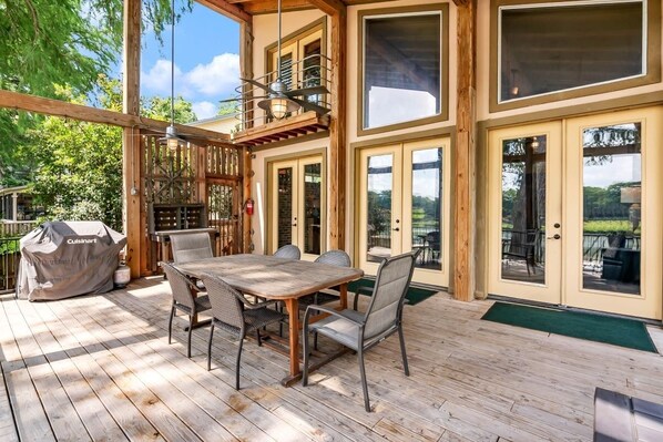 Una terraza de madera con una mesa y sillas de comedor, una parrilla cubierta y grandes puertas corredizas que dan a un patio.