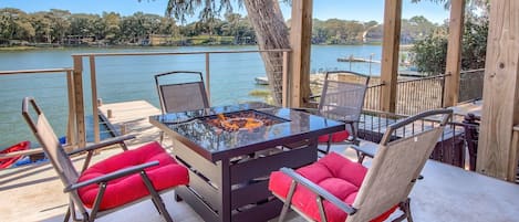 A glass-top table with red cushions, surrounded by metal-framed chairs, overlooking a lake with boats and trees.