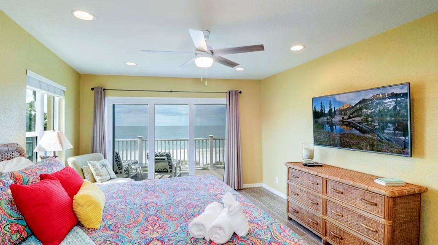 Habitación de hotel con una cama grande, ventilador de techo, vista al océano, zona de comedor con mesa y sillas, y un cuadro en la pared.