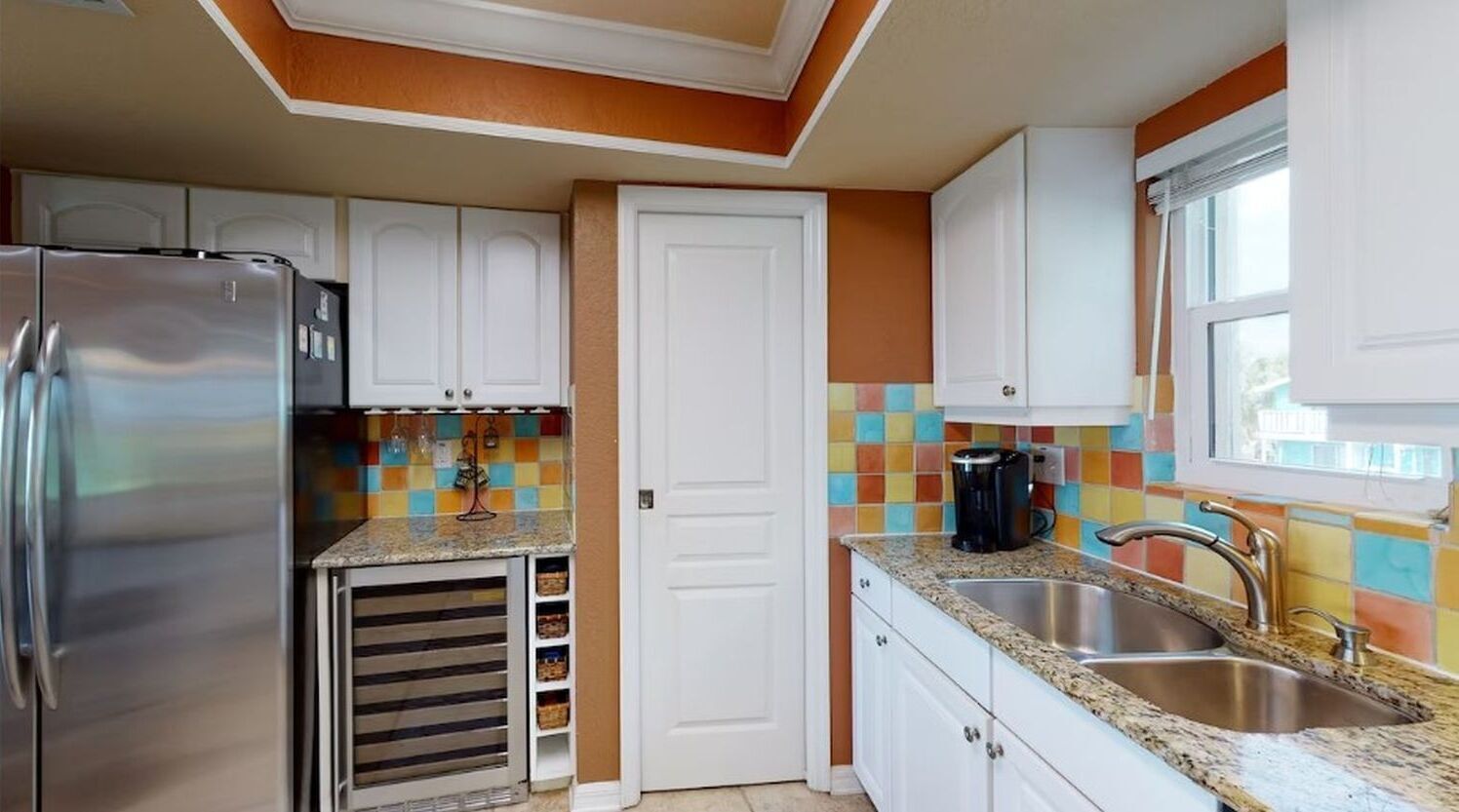 A kitchen with white cabinets, a stainless steel refrigerator, a colorful backsplash, and a window with blinds.