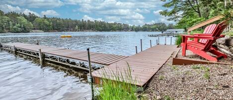Une jetée en bois s’étendant dans un lac paisible, entourée d’arbres, avec une chaise Adirondack rouge sur la droite.