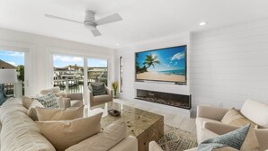 Een moderne woonkamer met een grote flatscreen-tv, een plafondventilator en uitzicht op de buitenomgeving door grote ramen.