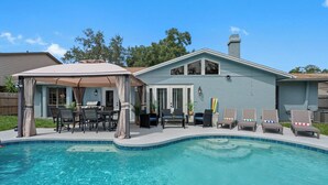 A backyard with a swimming pool, a covered patio area with outdoor furniture, and a house with a light blue exterior.