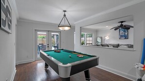 A pool table in a room with a ceiling fan, a window with a view of the outside, and a sofa against the wall.