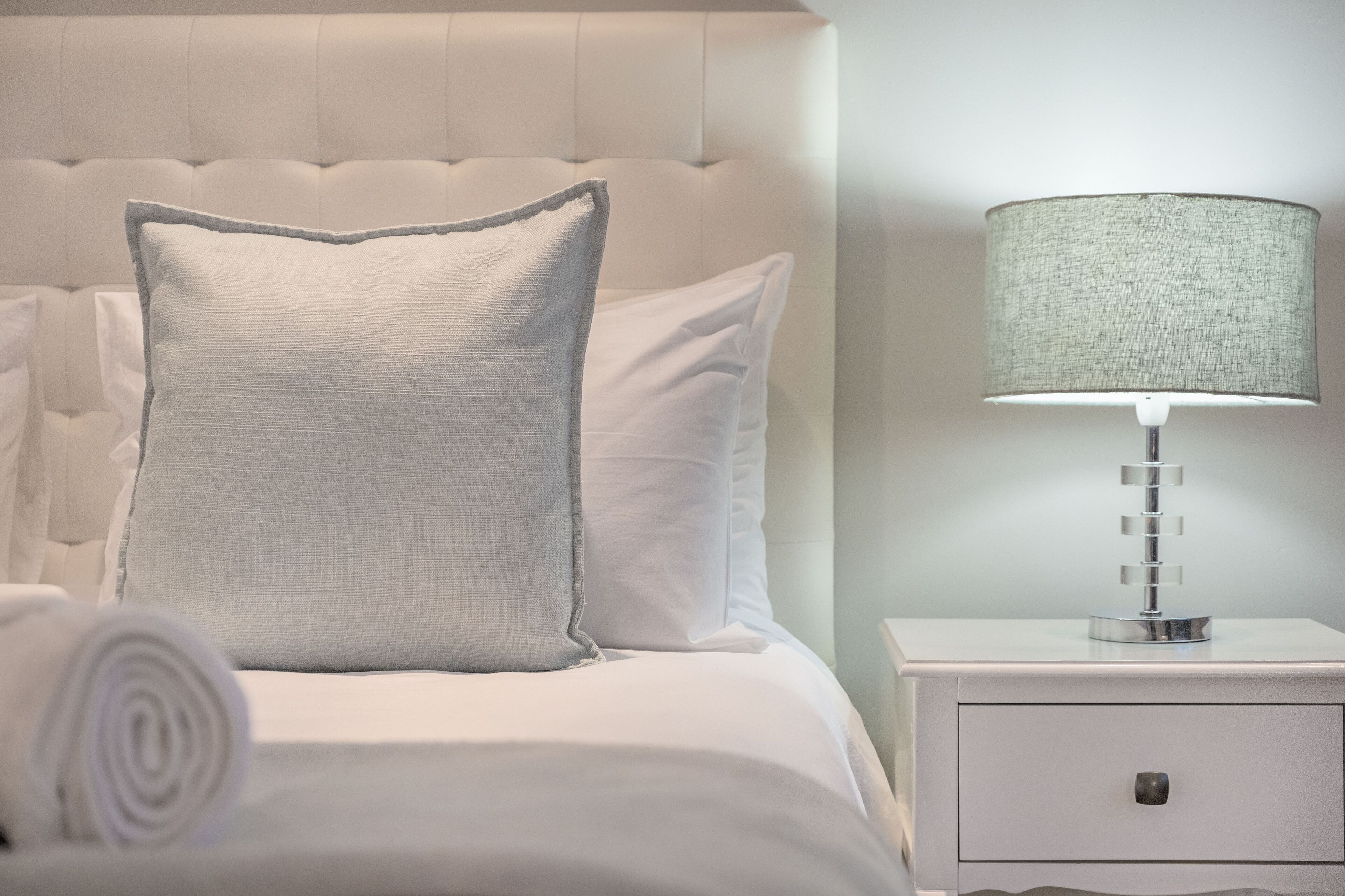 A neatly made bed with a pillow and a rolled towel, a bedside lamp, and a nightstand with a drawer.