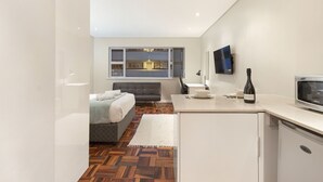 A modern hotel room with a bed, a small kitchenette, a flat-screen TV mounted on the wall, and a view of the city through the window. - Sandringham 55 (Cape Town)