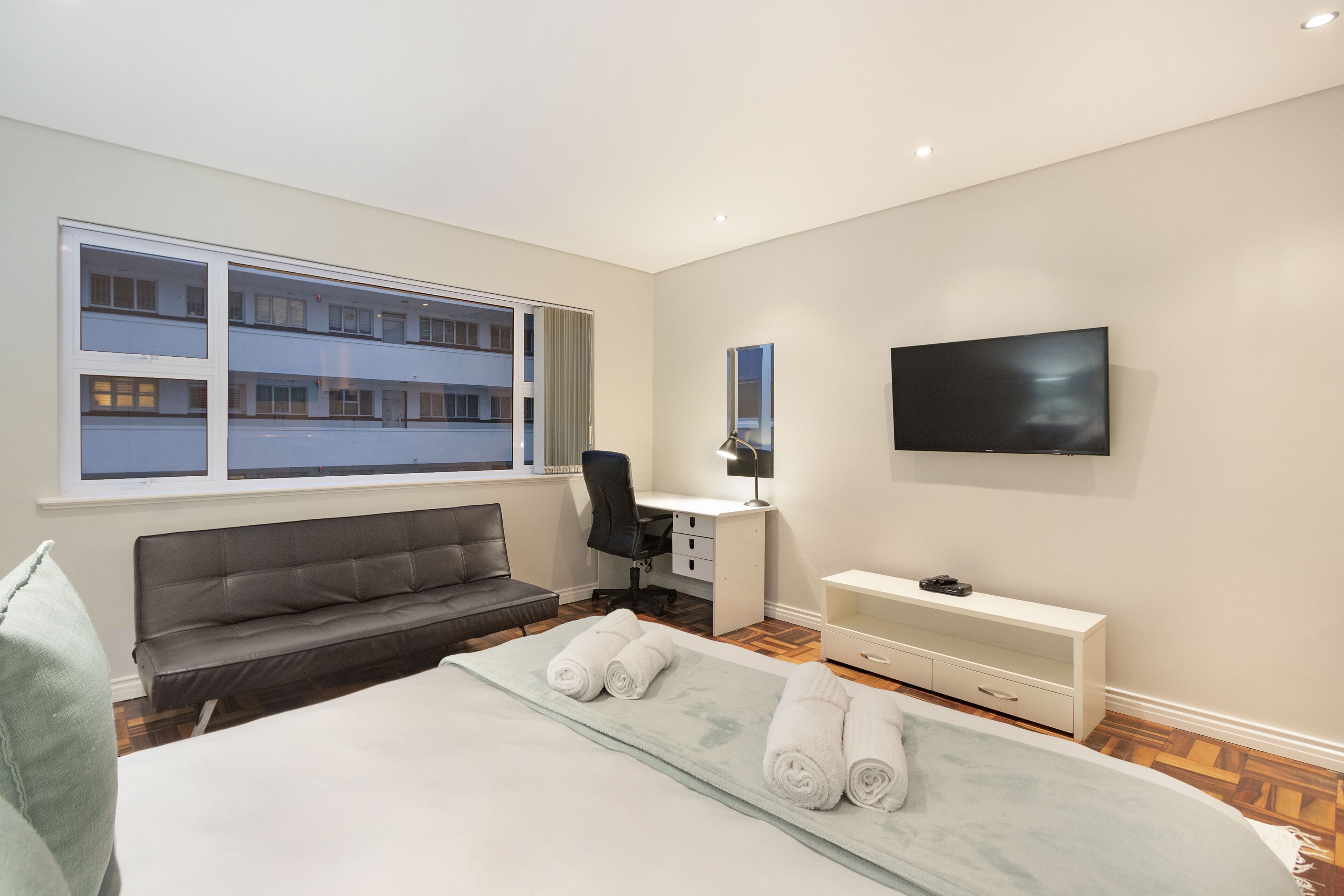 A modern hotel room with a large bed, a desk, a chair, a sofa, a TV, and a window with a view of other buildings.