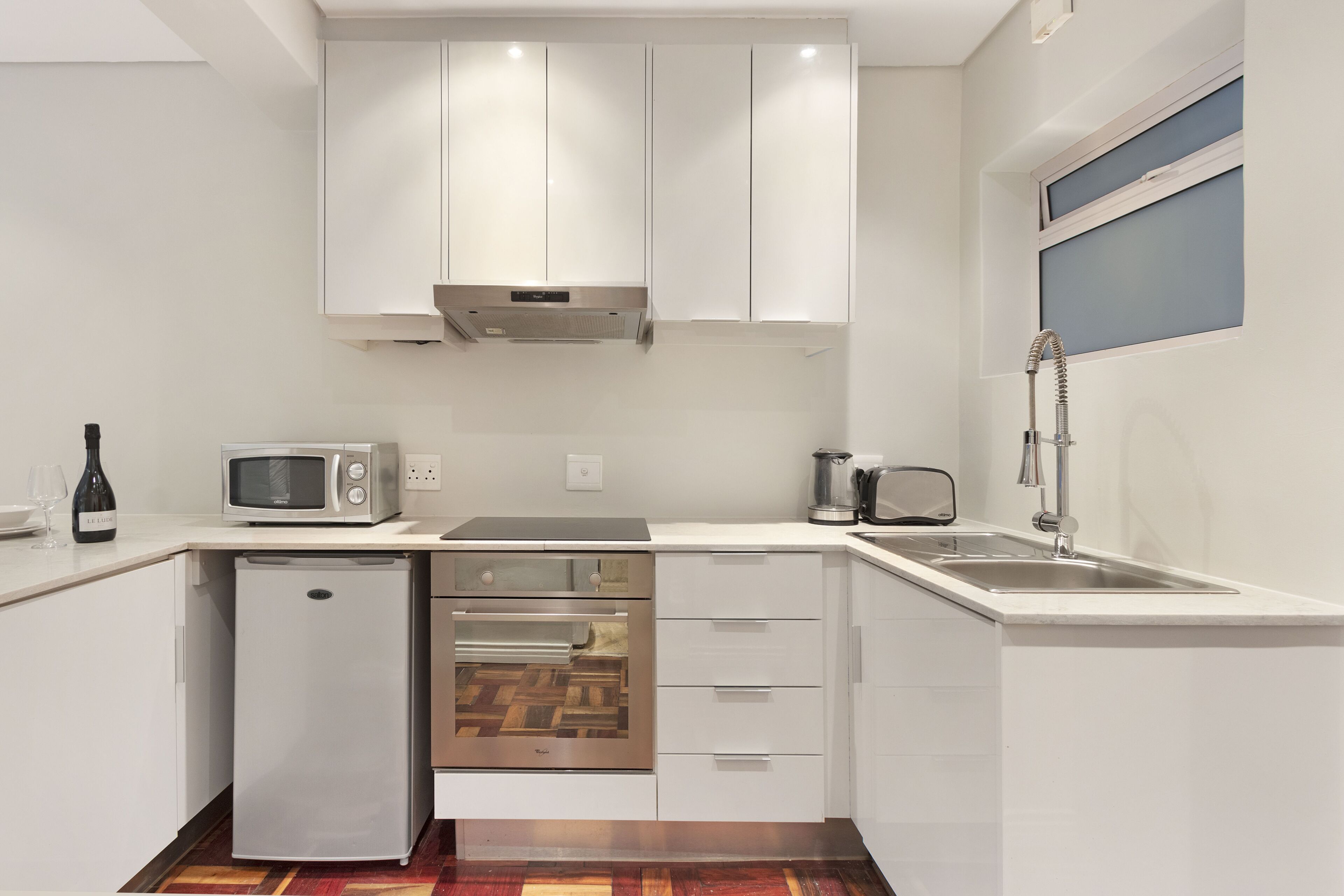Una cocina compacta con muebles blancos, un microondas integrado, un extractor de acero inoxidable, un refrigerador pequeño y un lavamanos con grifo.